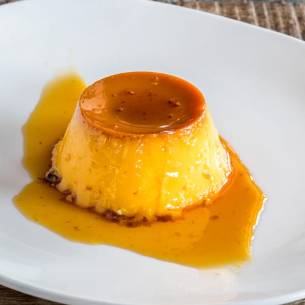 A golden flan dessert with caramel sauce on a white plate, against a rustic wooden background.