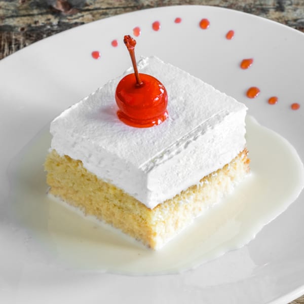 A slice of tres leches cake with whipped cream and a cherry on a white plate.
