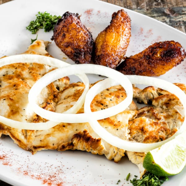 Grilled chicken with onion rings, fried plantains, parsley, and a lime wedge on a white plate.