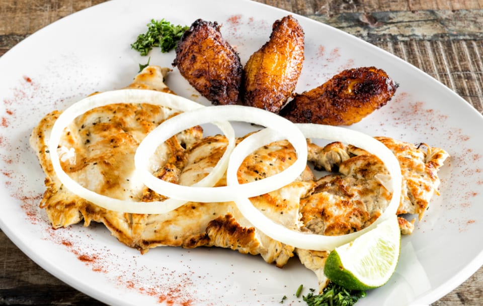 Grilled chicken with onion rings, fried plantains, parsley, and a lime wedge on a white plate.